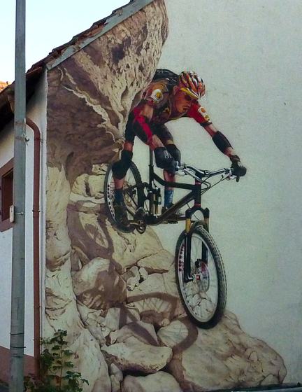 Hauswand mit Mountainbiker, der über einen gemalten Felsen springt